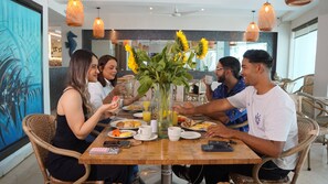 A group of people dining at a wooden table with a vase of sunflowers, surrounded by wicker chairs and orange pendant lights.