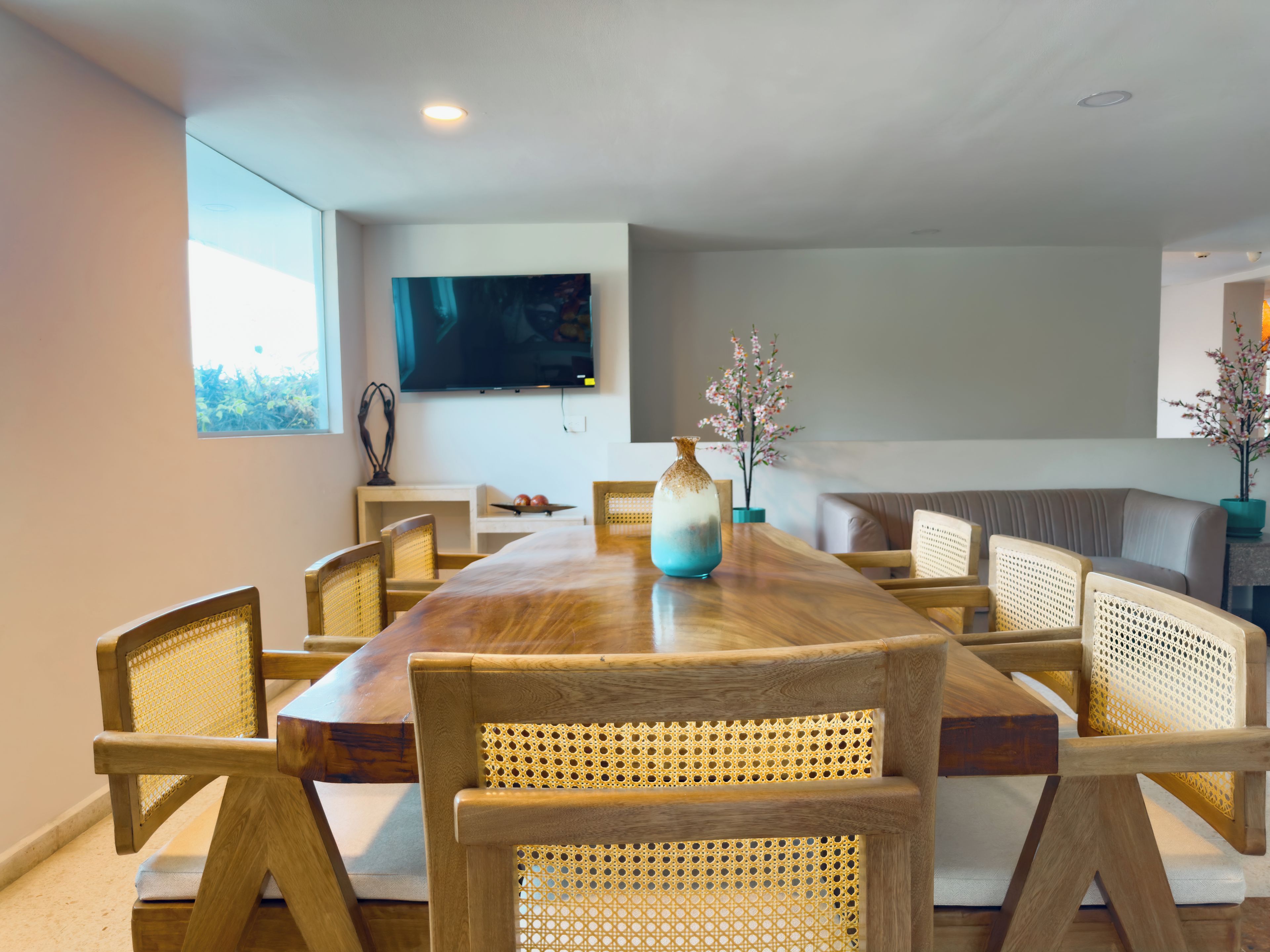 Un comedor con una larga mesa de madera, sillas de mimbre, un florero y un televisor de pantalla plana en la pared.