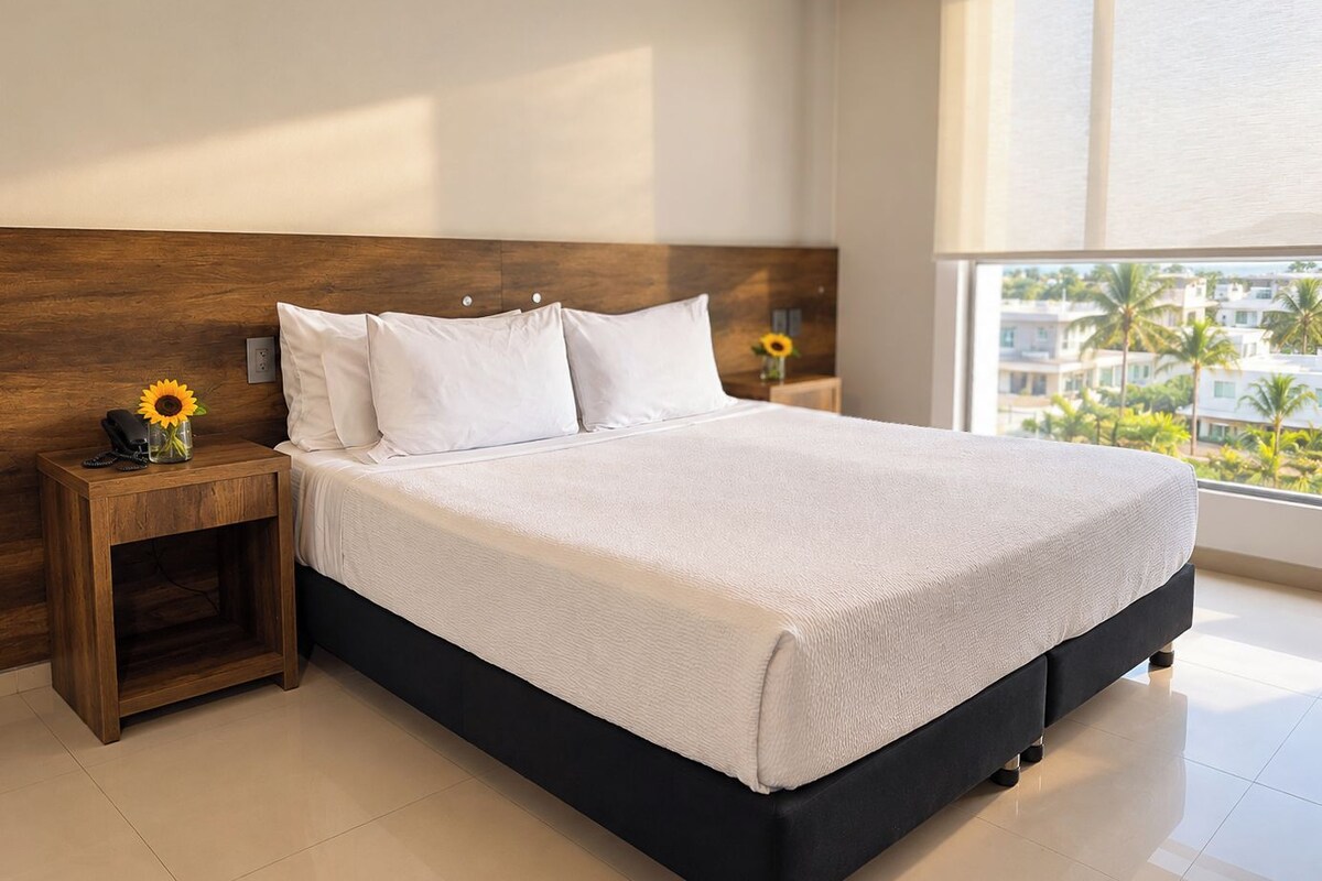A hotel room with a large bed, a wooden headboard, a nightstand with a vase of sunflowers, and a view of palm trees through the window.