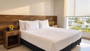 A hotel room with a large bed, a wooden headboard, a nightstand with a vase of sunflowers, and a view of palm trees through the window.