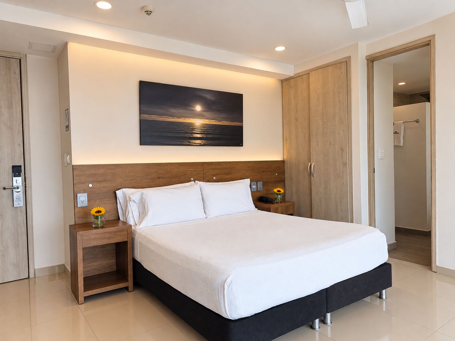 A modern hotel room with a large bed, wooden headboard, bedside tables with lamps, a painting of a sunset, and a door leading to a bathroom.