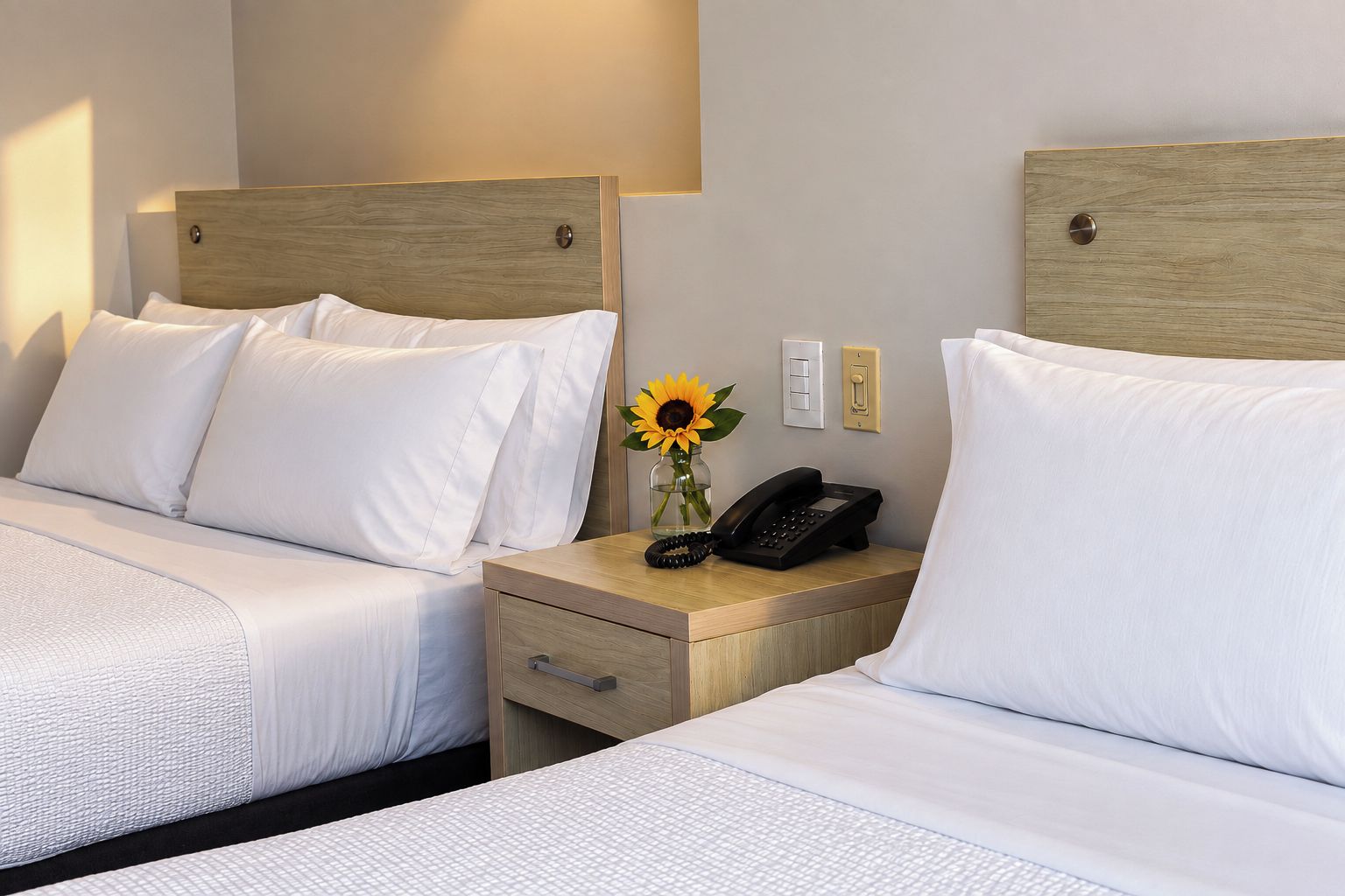 Une chambre d’hôtel avec deux lits, chacun doté de draps blancs et d’une tête de lit en bois. On y trouve une table de chevet avec un téléphone et un vase contenant un tournesol.