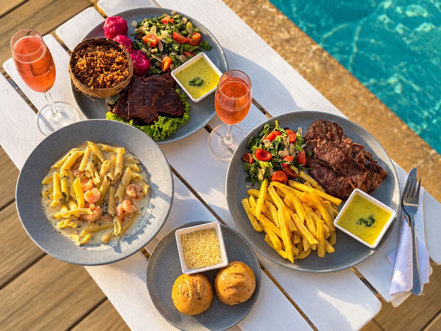 A dining table with plates of food, glasses of beverages, and a poolside setting.