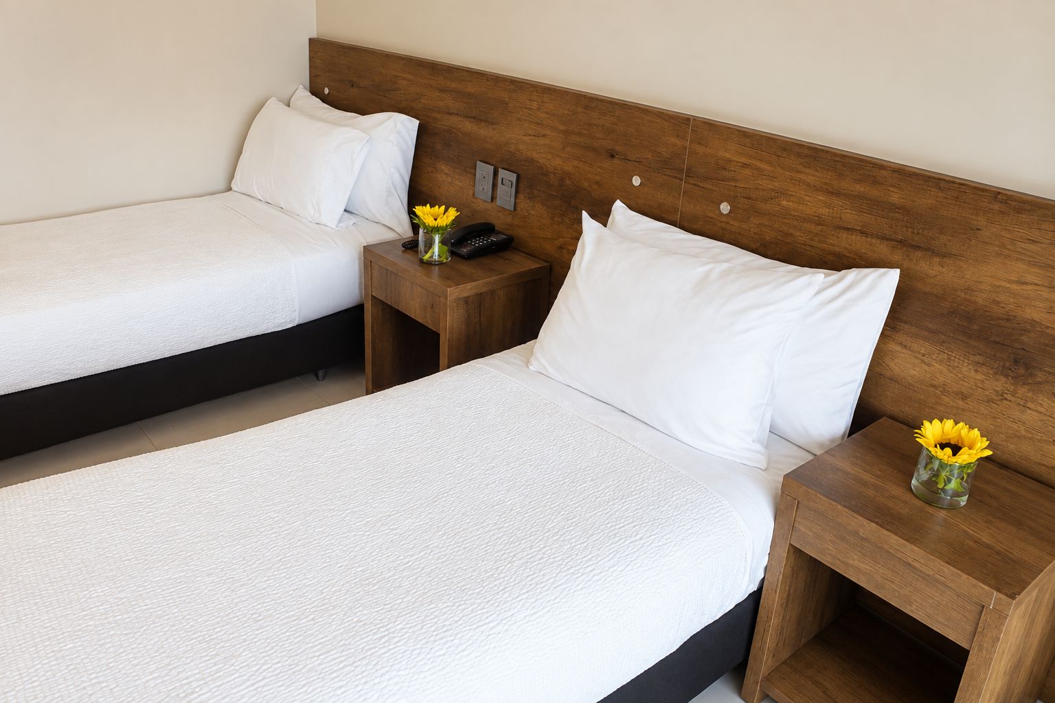 Una camera d'albergo con due letti singoli, poggiatesta in legno, comodini con lampade e un vaso con fiori gialli su ciascun tavolo.