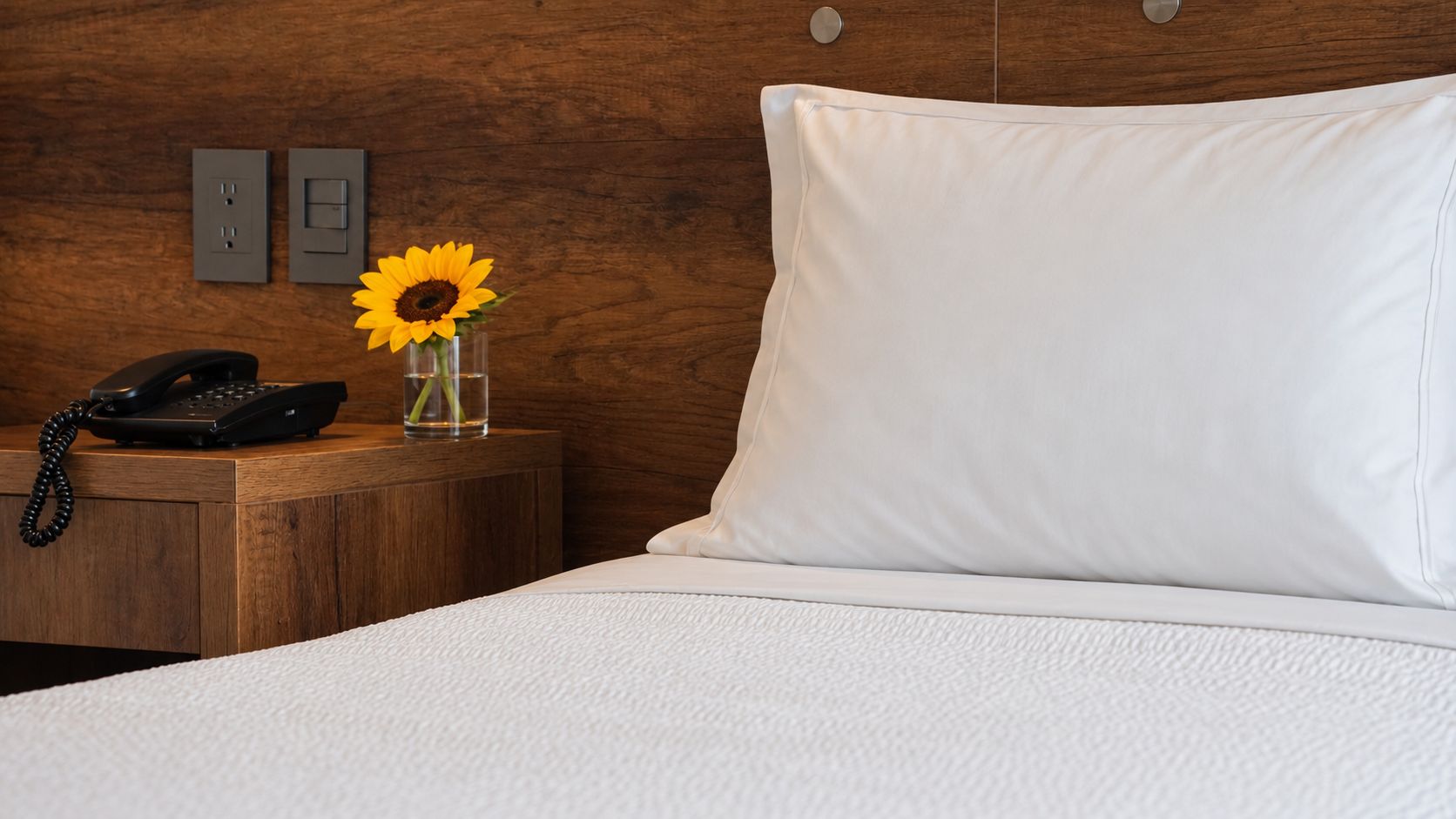 Habitación de hotel con cabecera de madera, cama blanca, mesita de noche con teléfono y un jarrón con un girasol.
