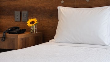 Habitación de hotel con cabecera de madera, cama blanca, mesita de noche con teléfono y un jarrón con un girasol.