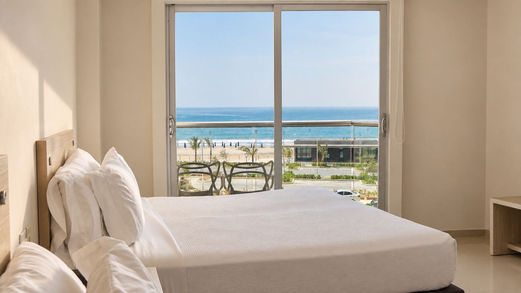 Habitación de hotel con una cama grande, vista a la playa y un balcón con mobiliario exterior.