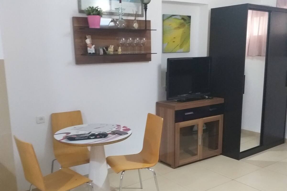 A dining area with a round table and four chairs, a television, a cabinet, and a shelf with decorative items.