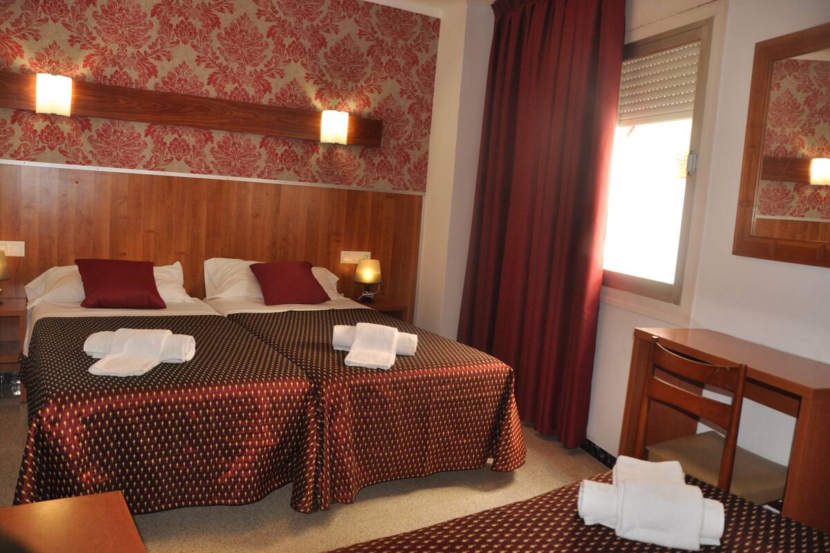 Une chambre d’hôtel avec deux lits, une tête de lit en bois, un bureau et une fenêtre avec des rideaux.