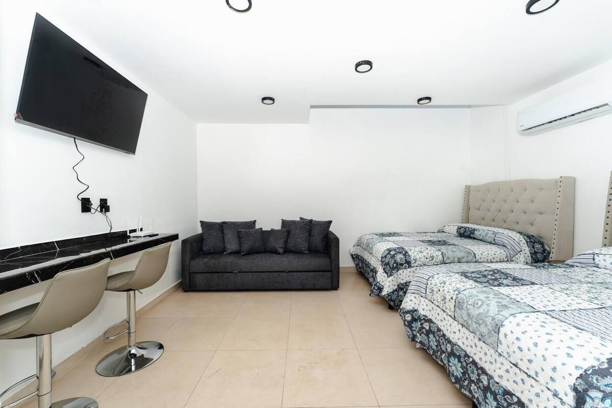 A modern bedroom with a sofa, a bed with patterned bedding, a TV mounted on the wall, and a bar area with stools.