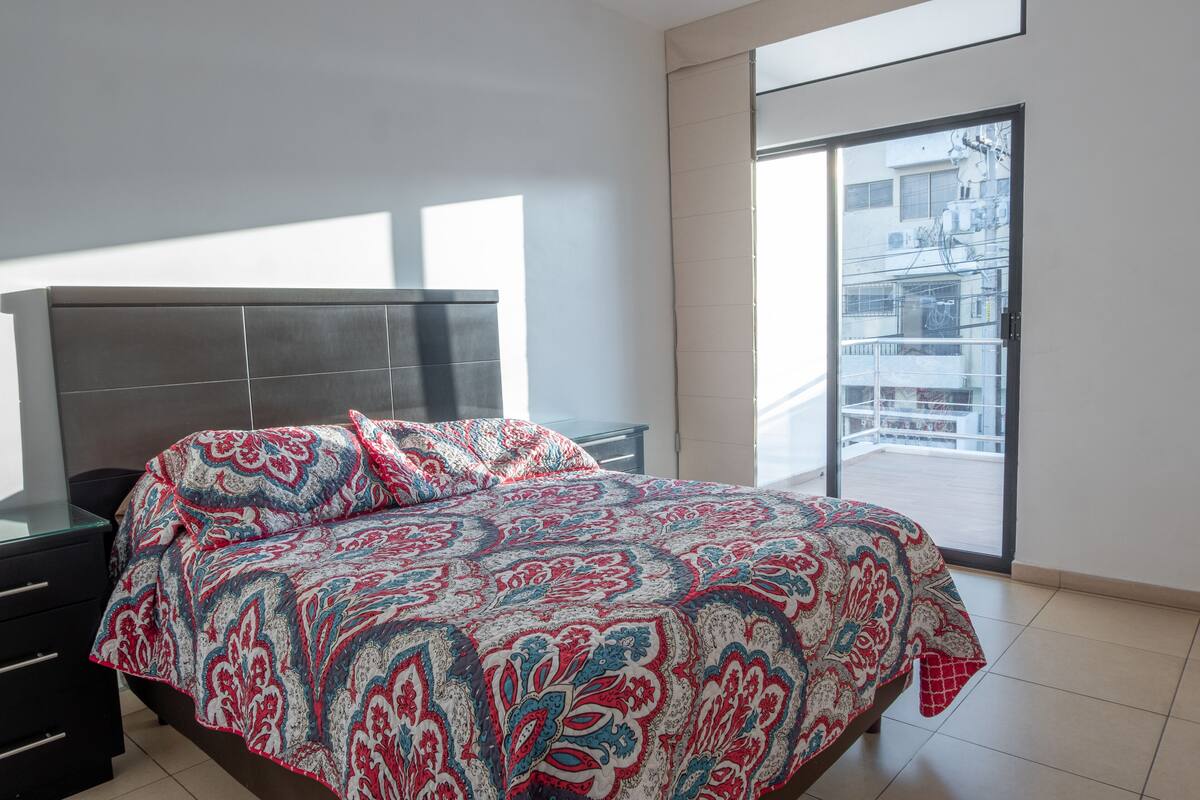 A modern bedroom with a large bed, a glass desk, and a balcony with a view of buildings.
