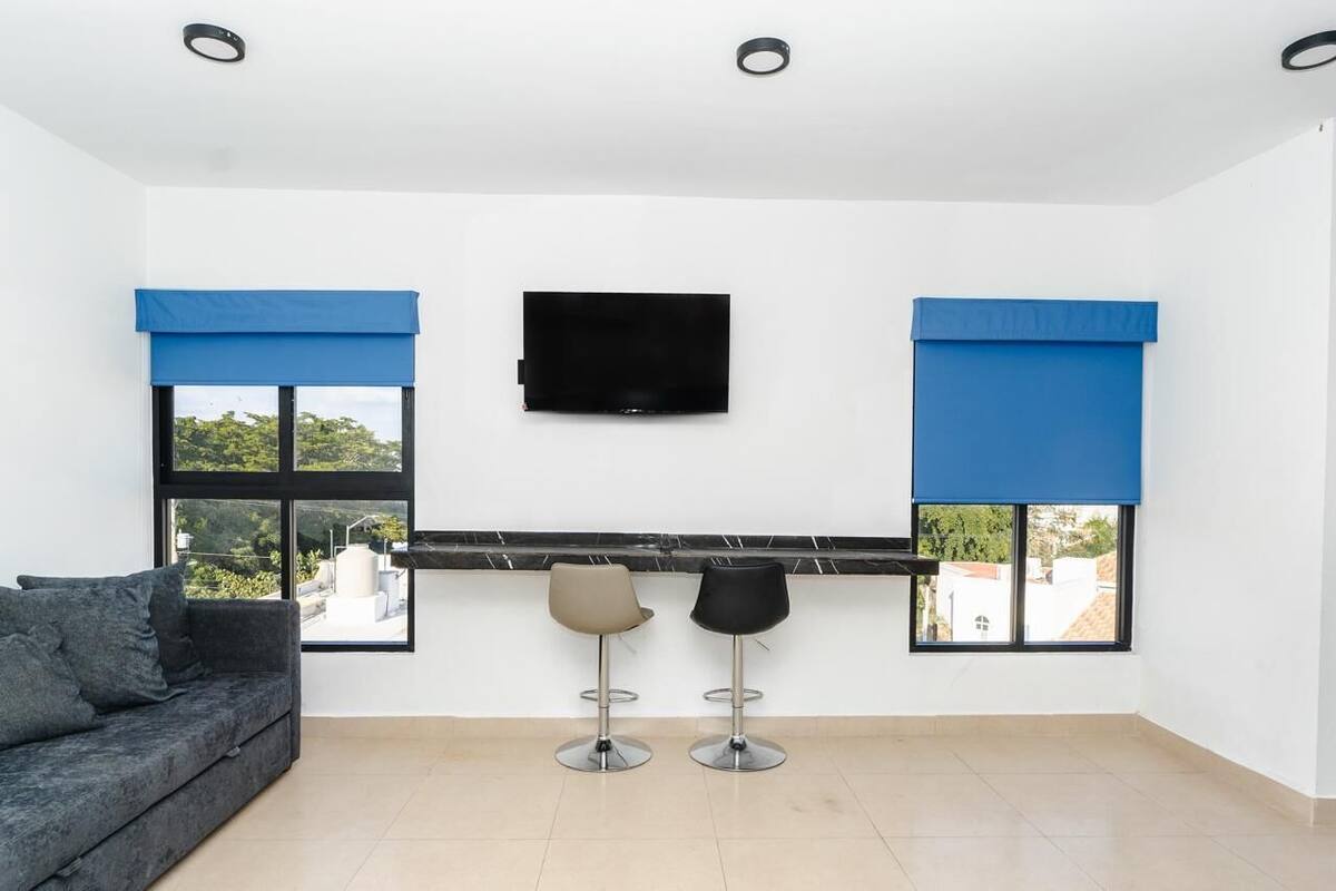 A modern room with a sofa, two bar stools, a flat-screen TV, and large windows with blue blinds.
