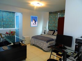 Quarto com uma cama, uma escrivaninha com computador, uma cadeira, uma janela com cortinas e um quadro na parede.