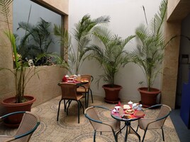 Uma área de refeições com cadeiras de vime e uma mesa posta para o café da manhã, cercada por plantas em vasos.