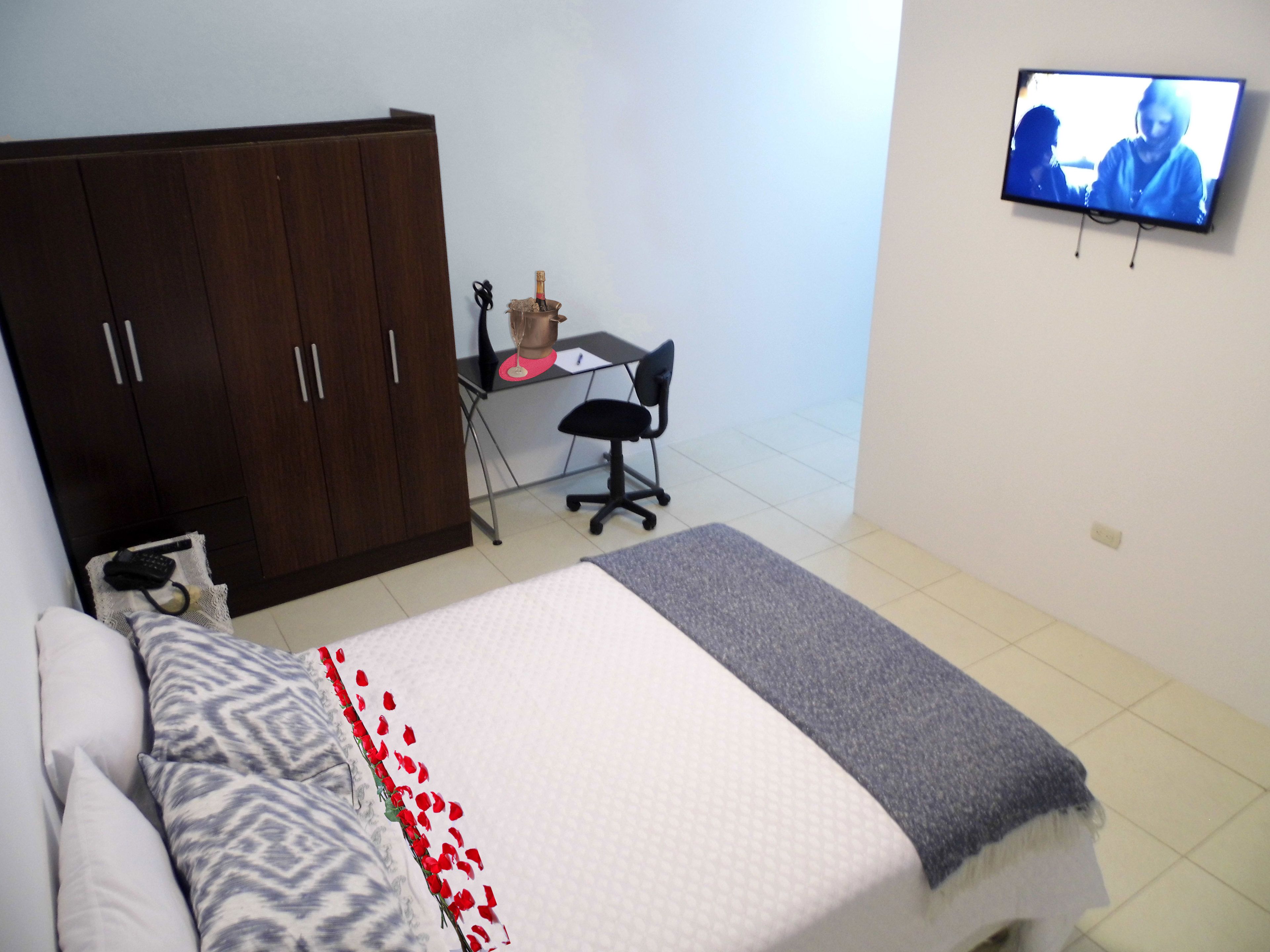 A bedroom with a bed, a wardrobe, a desk with a chair, and a television on the wall.