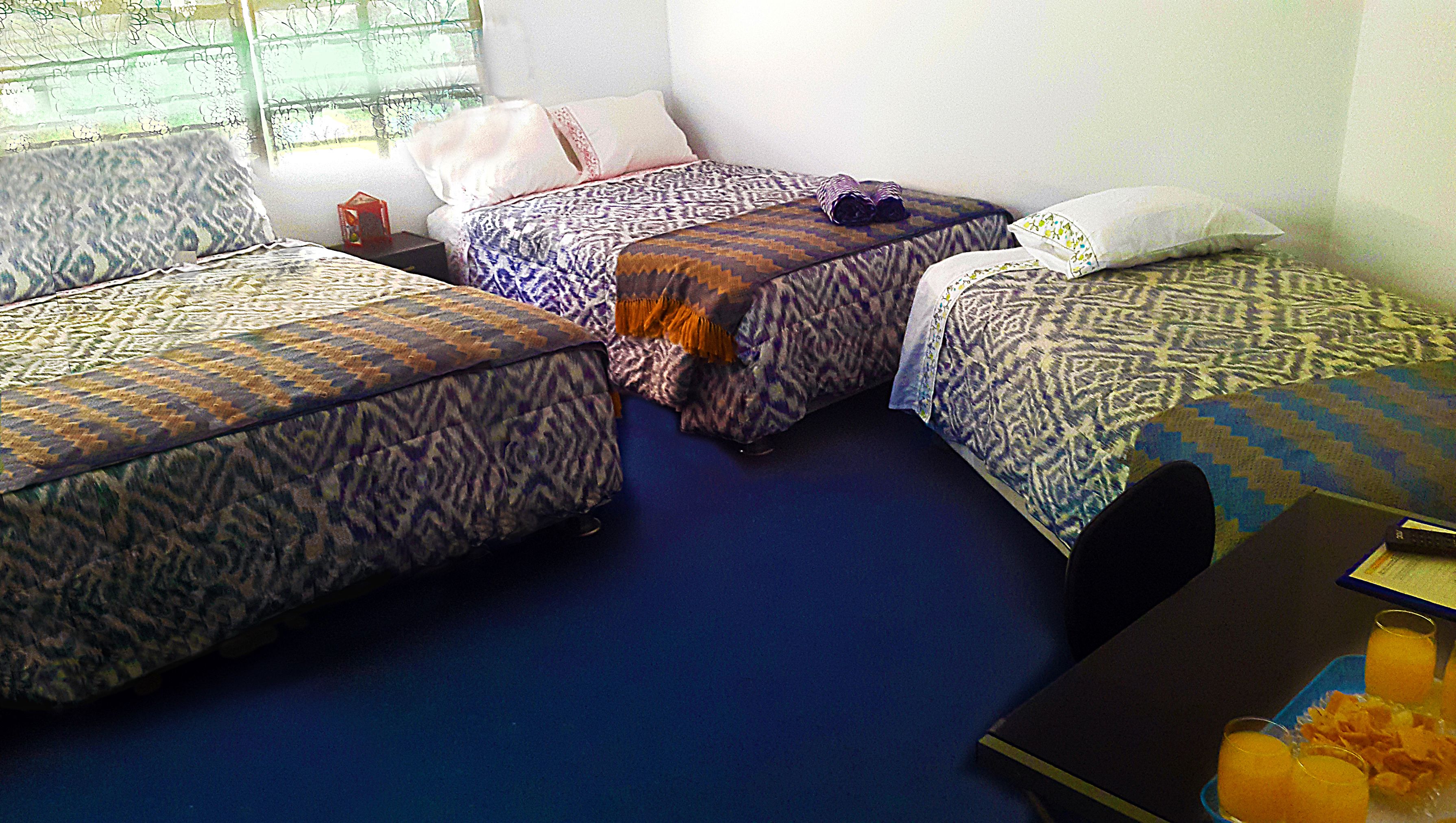 Ein Zimmer mit drei Betten, alle mit gemusterten Bettbezügen. Es gibt einen kleinen Tisch mit einem Buch und Gläsern, sowie ein Fenster mit Vorhängen.