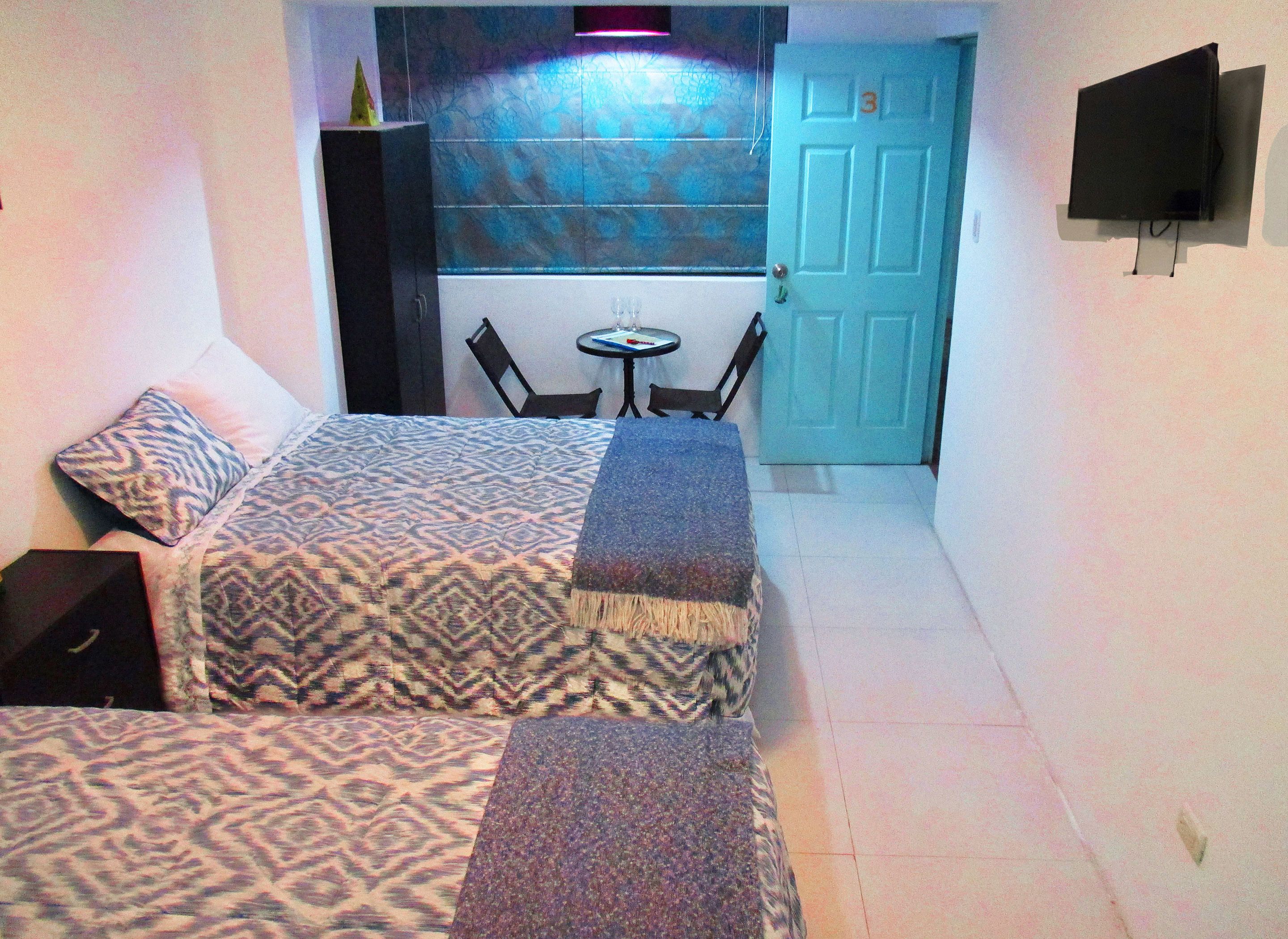 A small, compact room with two beds, a small table, and a TV mounted on the wall.