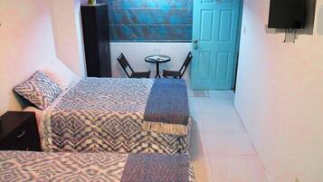 A small, compact room with two beds, a small table, and a TV mounted on the wall.