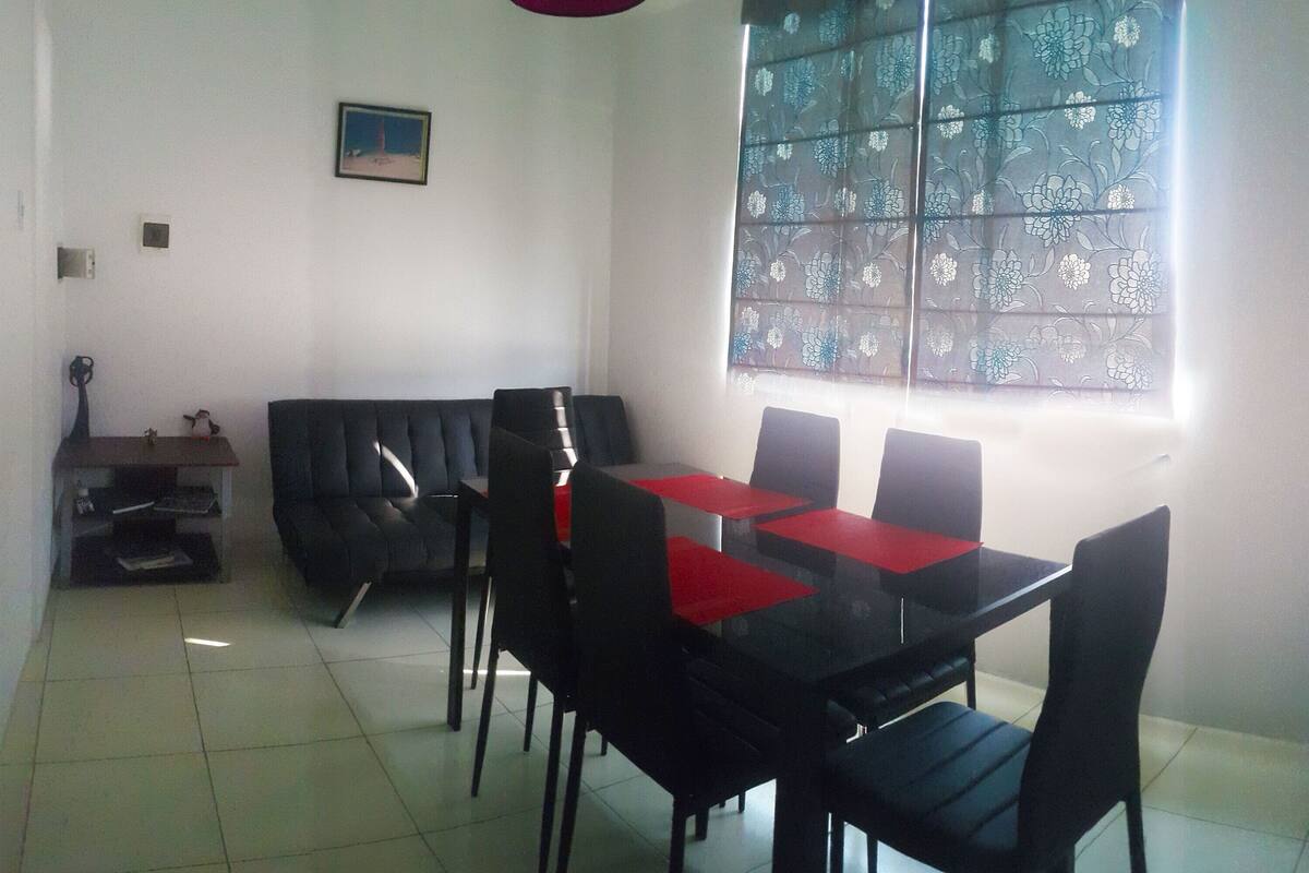Un coin repas avec une table et des chaises noires, un chemin de table rouge, un canapé noir et une fenêtre avec des stores à motifs floraux.