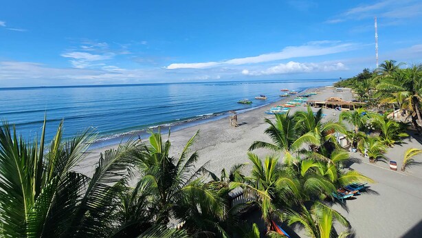 A beach with clear blue water, palm trees, and a few boats. - Bakasyunan Resort Zambales (Iba)