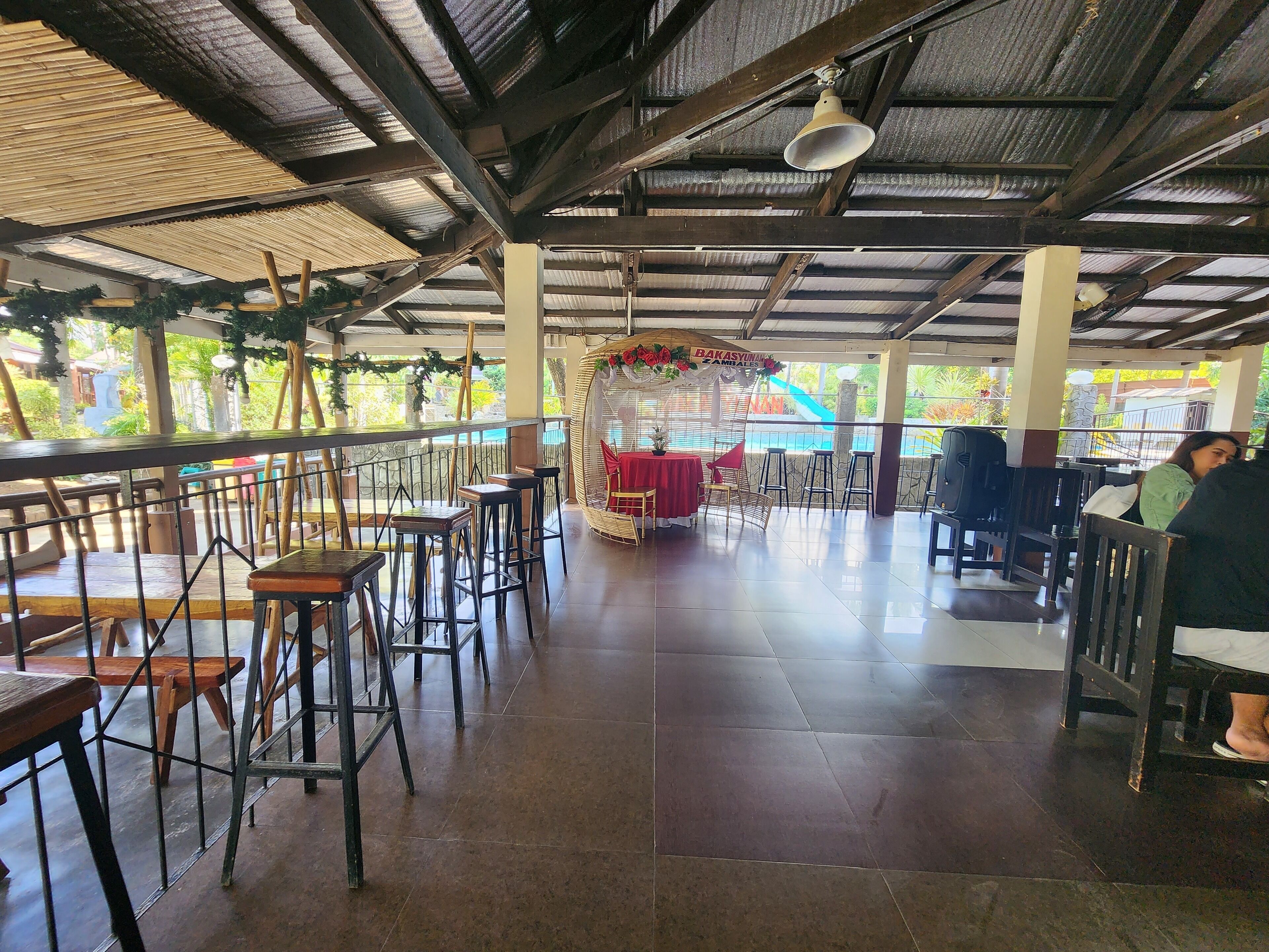 An indoor dining area with high ceilings, metal beams, and a tiled floor. There are several tables and chairs, some with red tablecloths. A decorative arch with greenery is in the background.