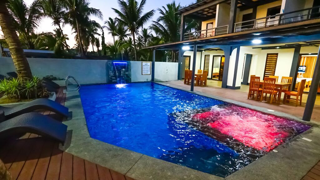 Une piscine agrémentée d’un élément décoratif aquatique, entourée de palmiers et bordée d’un bâtiment doté d’une terrasse.