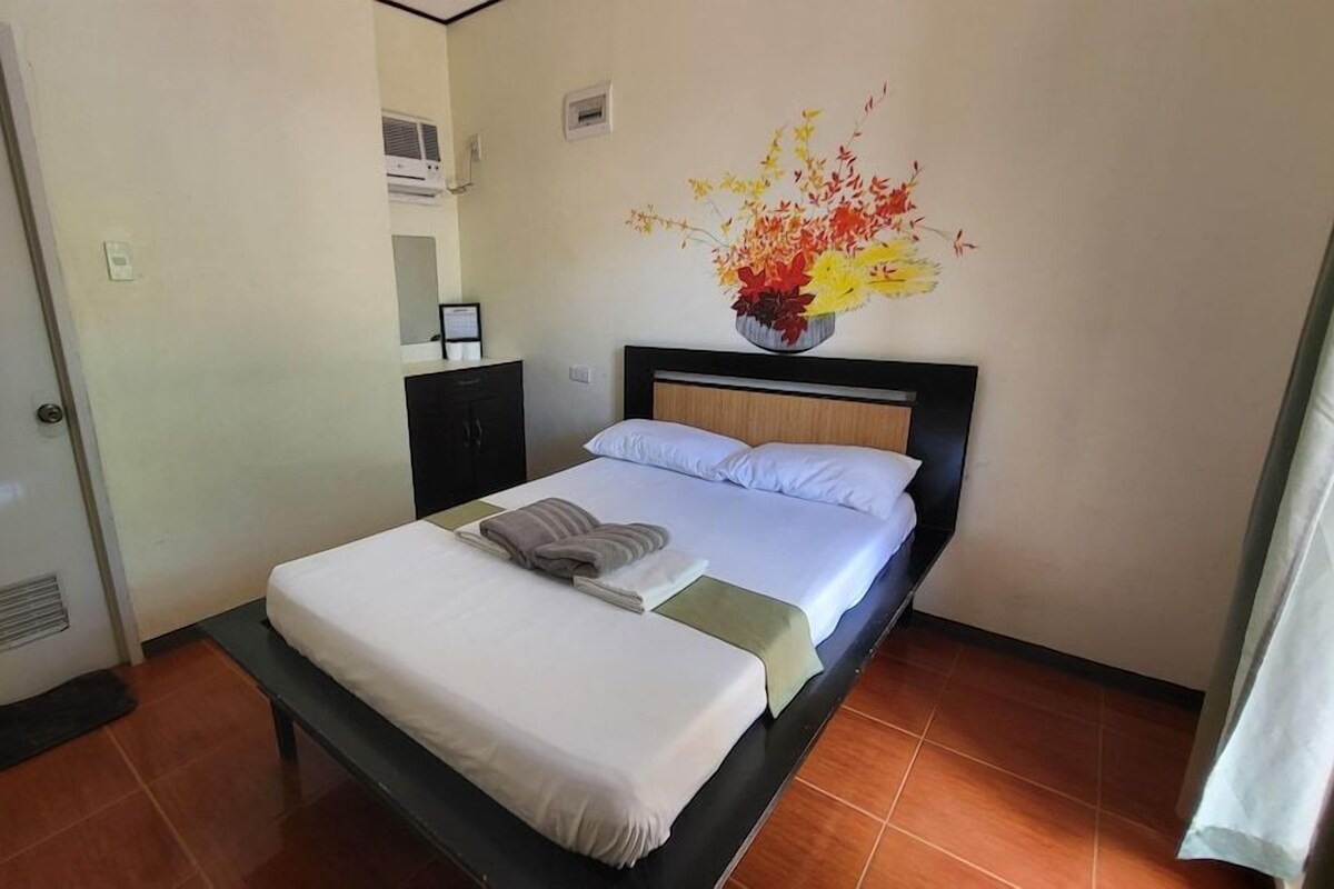 A hotel room with a bed, a nightstand, a wall-mounted air conditioner, and a decorative vase with flowers.