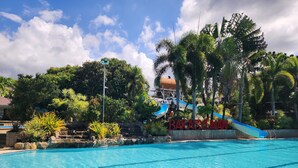 A swimming pool with a water slide, surrounded by tropical vegetation and a clear sky. - Bakasyunan Resort Zambales (Iba)