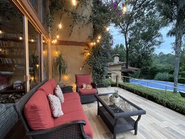 Un espacio exterior acogedor con mobiliario de mimbre e iluminado con luces colgantes, con vistas a una piscina y abundante vegetación.