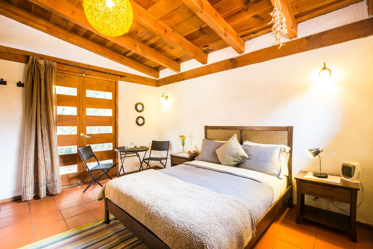A cozy bedroom with a wooden ceiling, a bed with pillows, a nightstand with a lamp, and a chair by the window.