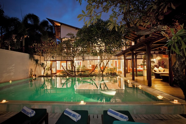 A swimming pool with lounge chairs, a wooden deck, and a covered seating area with plants.