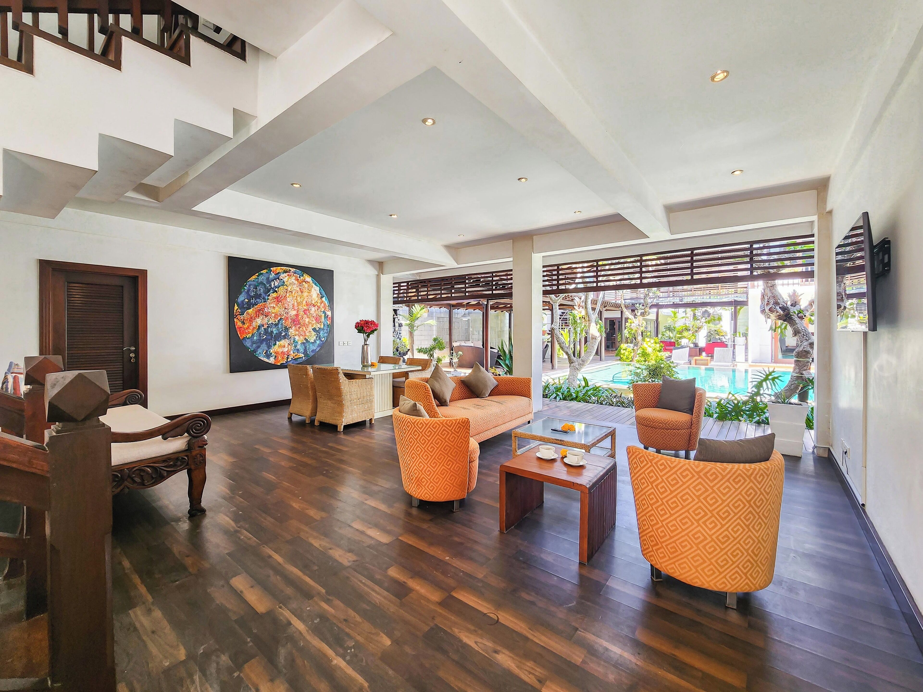 A spacious living area with a wooden floor, orange patterned sofas, a dining table, and a large painting on the wall.