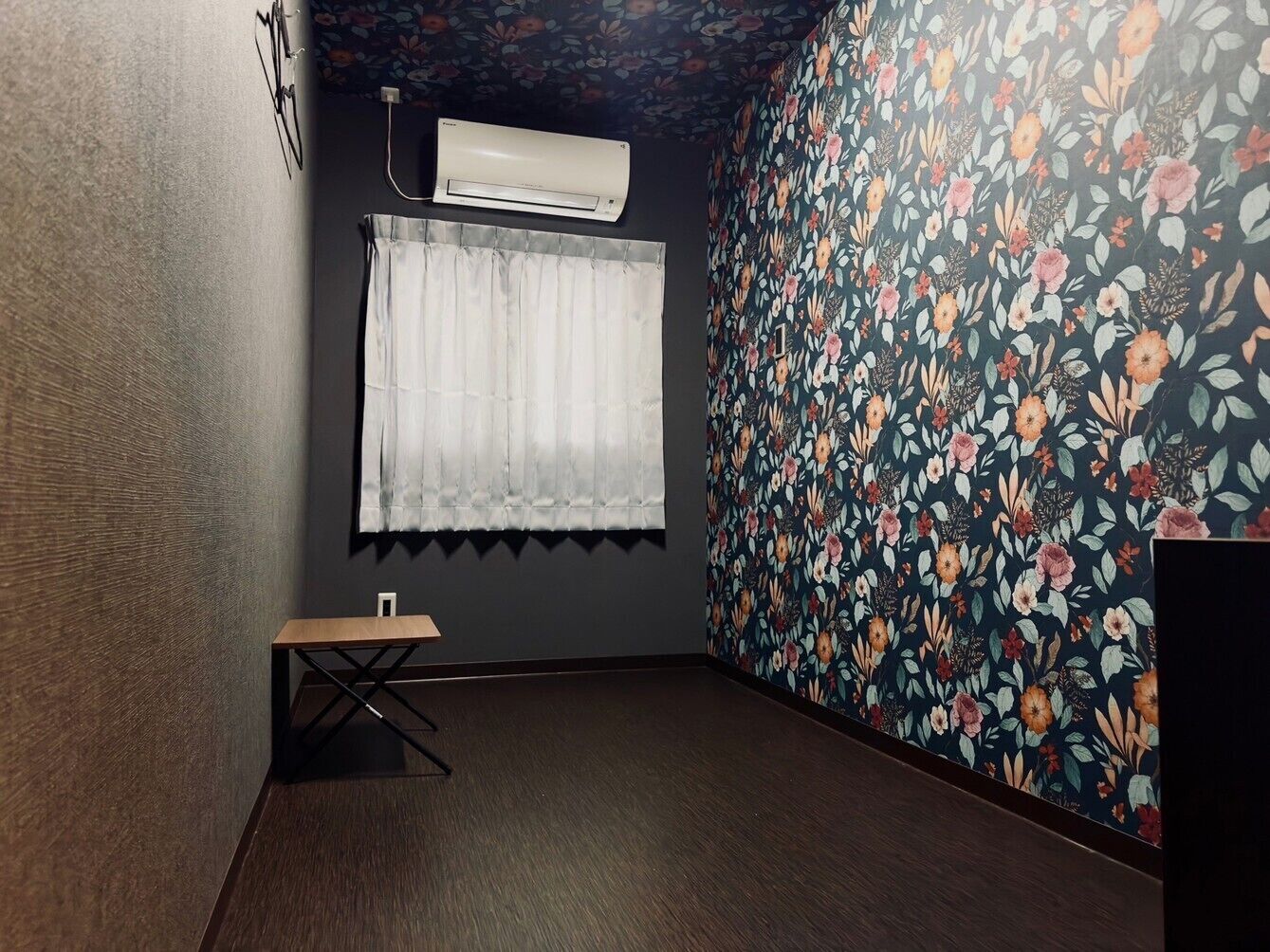 A room with a dark wooden floor, a small table, a white curtain, and a floral wallpaper.