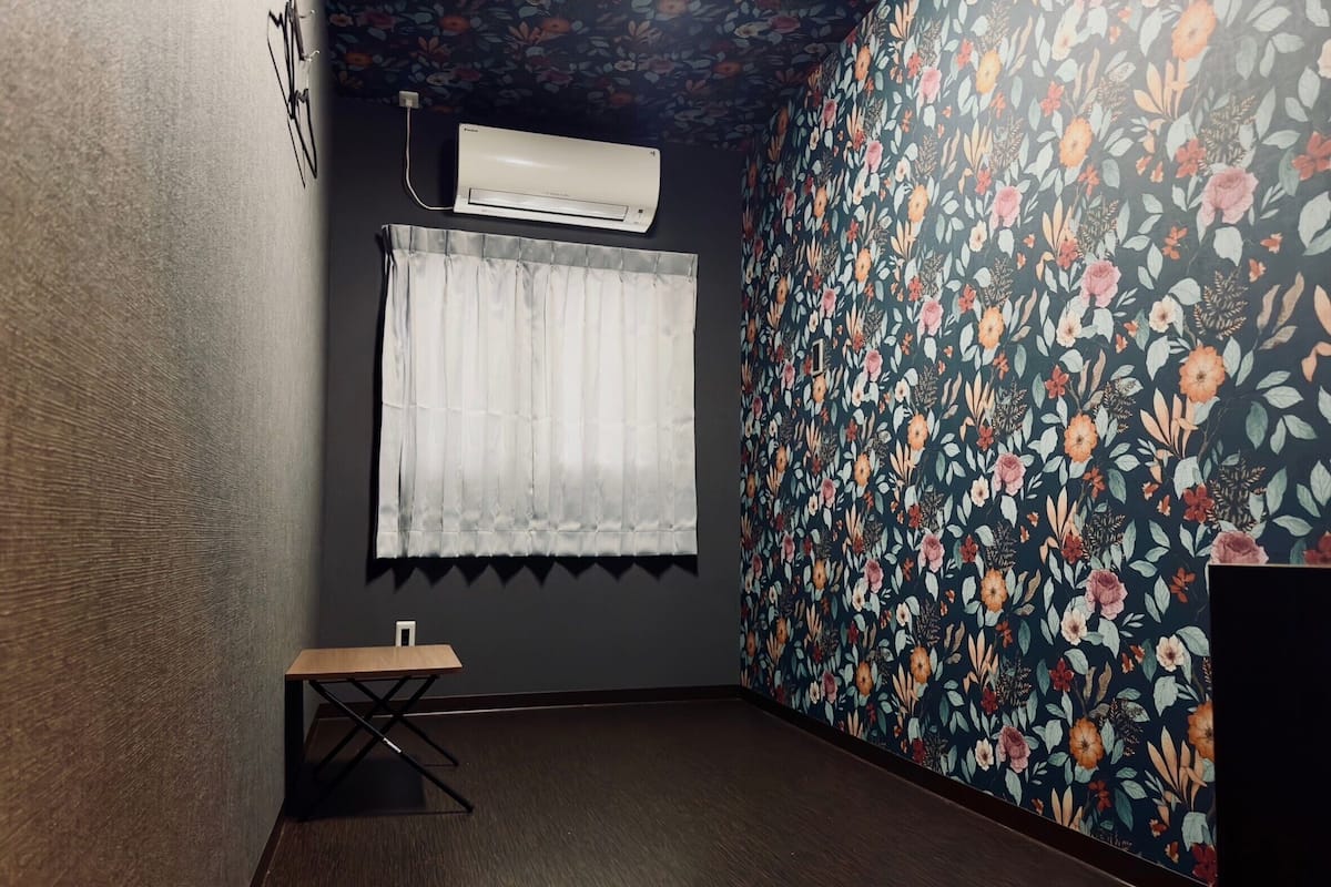 Ein Zimmer mit dunklem Holzboden, einer weißen Wandklimaanlage, einem kleinen Holztisch und einem Fenster mit weißem Vorhang.