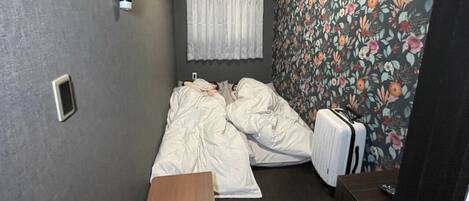 A small, compact room with two beds, a wooden desk, a suitcase, and a wall-mounted air conditioner.