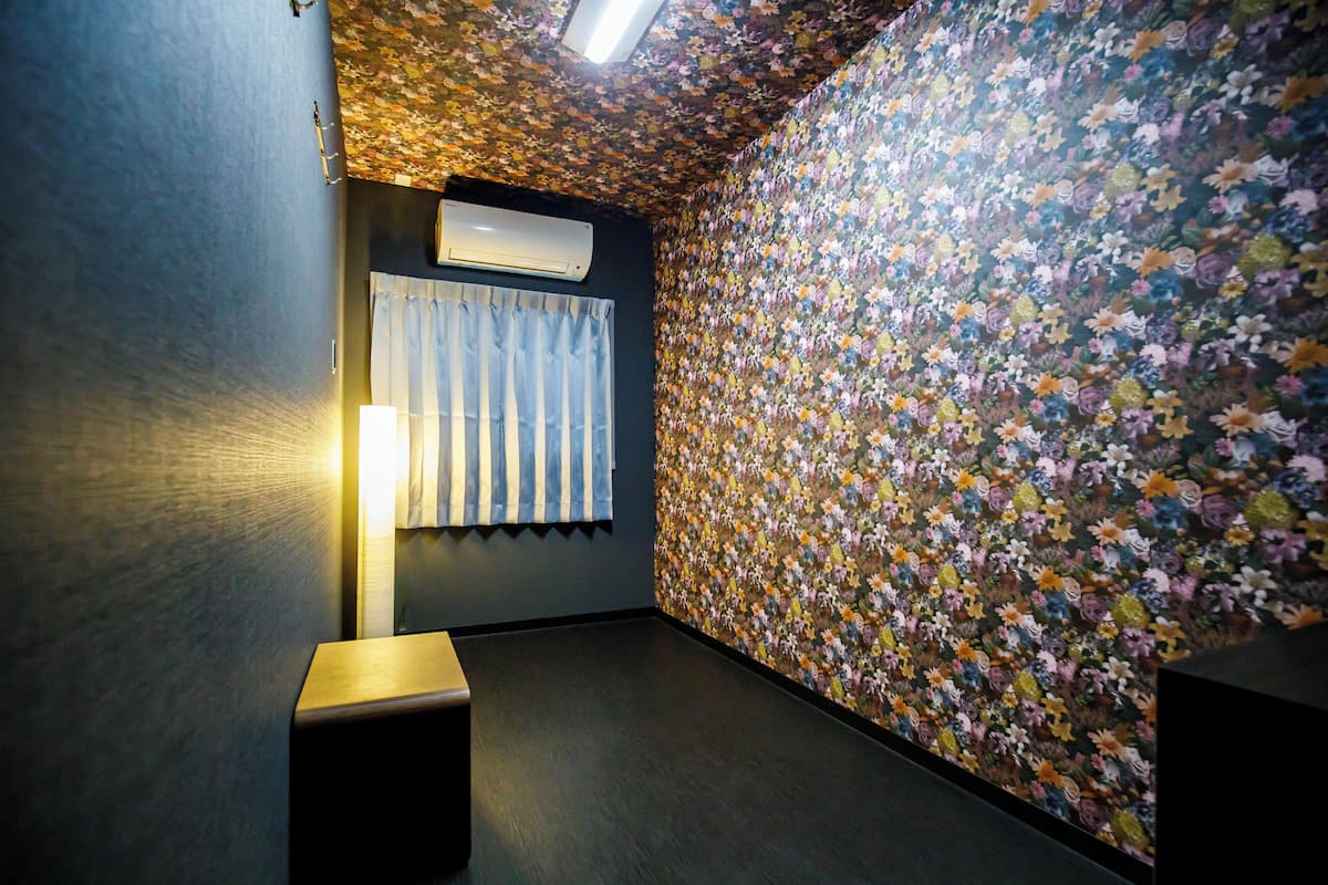 Ein Zimmer mit Blumenmuster-Tapete, Deckenventilator, Fenster mit Vorhängen und einer Stehlampe.