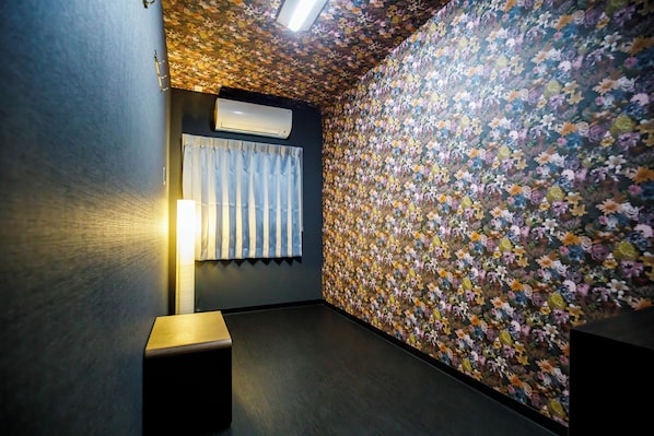 Een kamer met bloemen behang, een plafondventilator, een raam met gordijnen en een staande lamp.