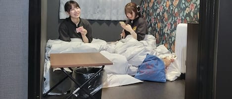 Deux personnes sont assises sur un lit, dans une pièce dont les murs sont recouverts d’un papier peint à motifs floraux. On y voit également une petite table et une poubelle.