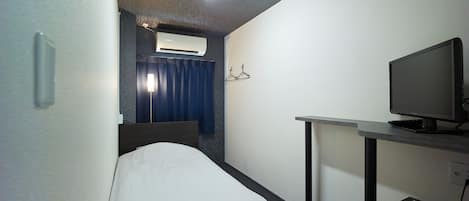 Ein kleines Hotelzimmer mit einem Einzelbett, einem Schreibtisch mit Computer, einem Fernseher und einer Klimaanlage.