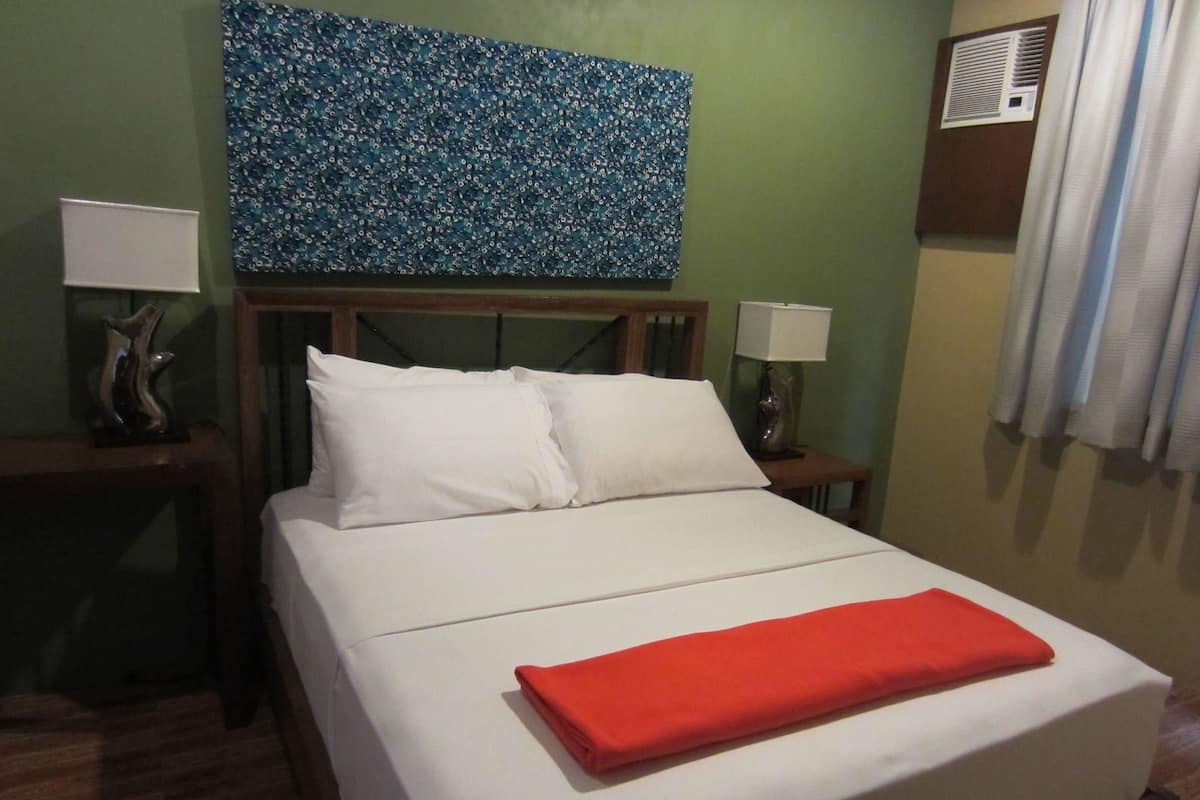 Quarto de hotel com uma cama, duas mesas de cabeceira, um criado-mudo, ar condicionado fixado na parede e uma janela com cortinas.
