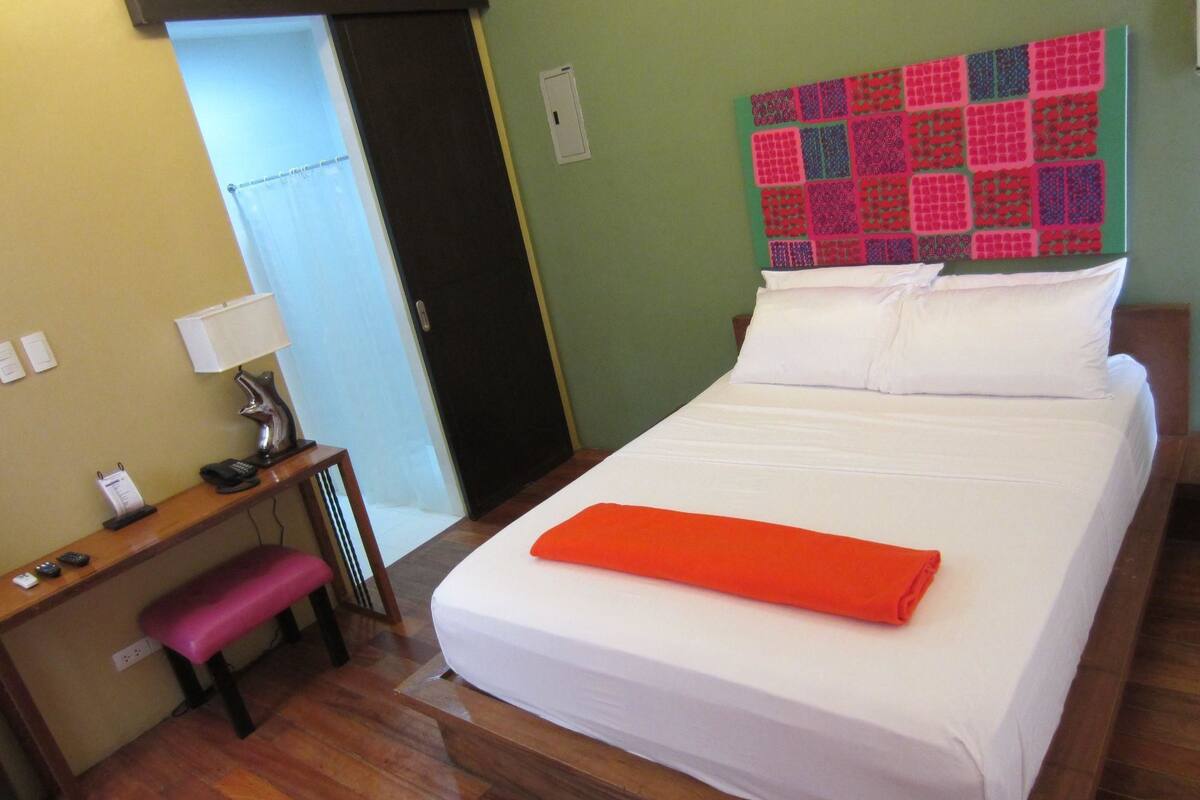 Quarto de hotel com uma cama, uma mesa de trabalho, um banco e uma decoração de parede colorida.