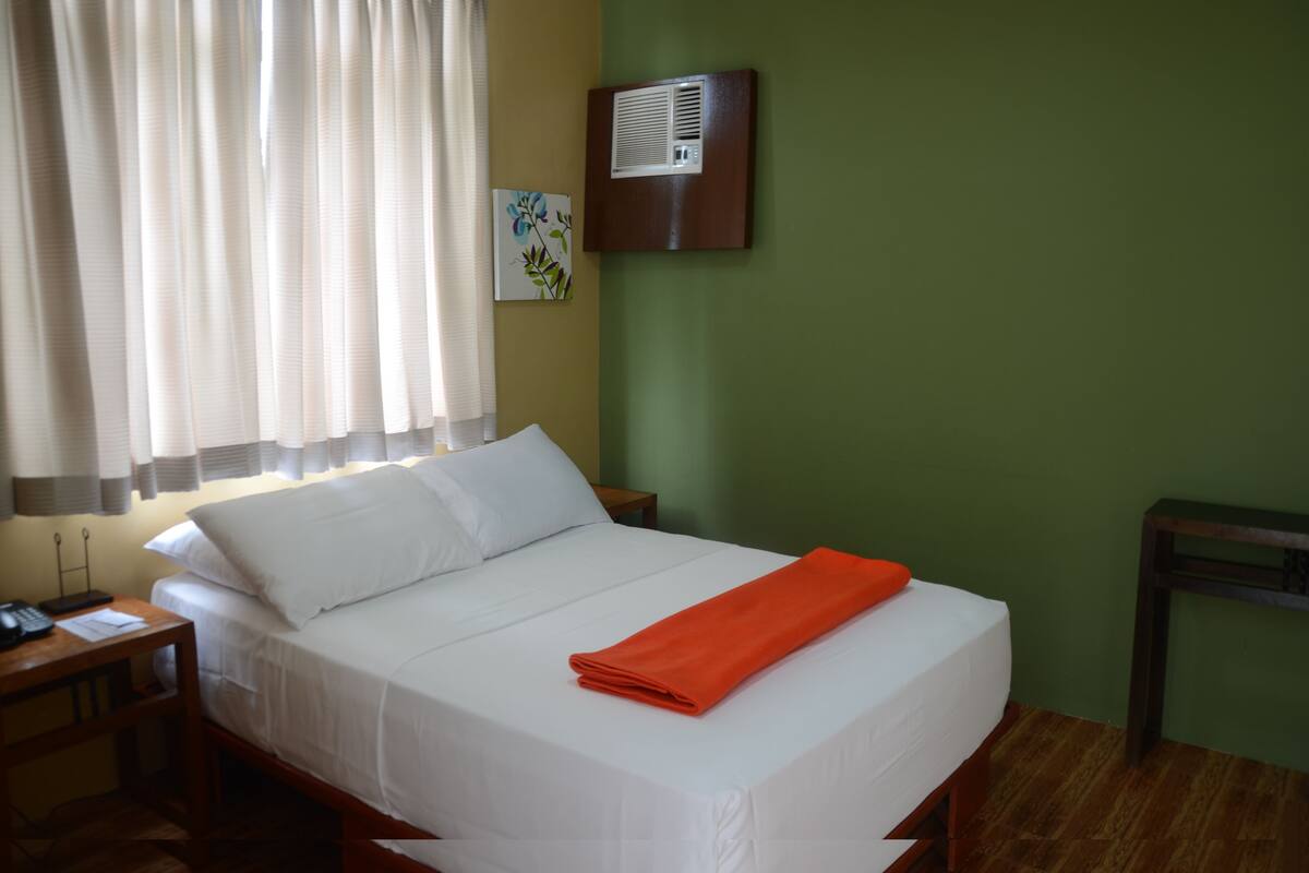 Uma cama bem arrumada com roupa de cama branca e uma toalha laranja, uma mesa de cabeceira com abajur, um ar condicionado fixado na parede e um quadro na parede.
