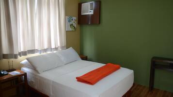 Una cama bien hecha con sábanas blancas y una toalla naranja, una mesita de noche con una lámpara, un aire acondicionado de pared y un cuadro en la pared.