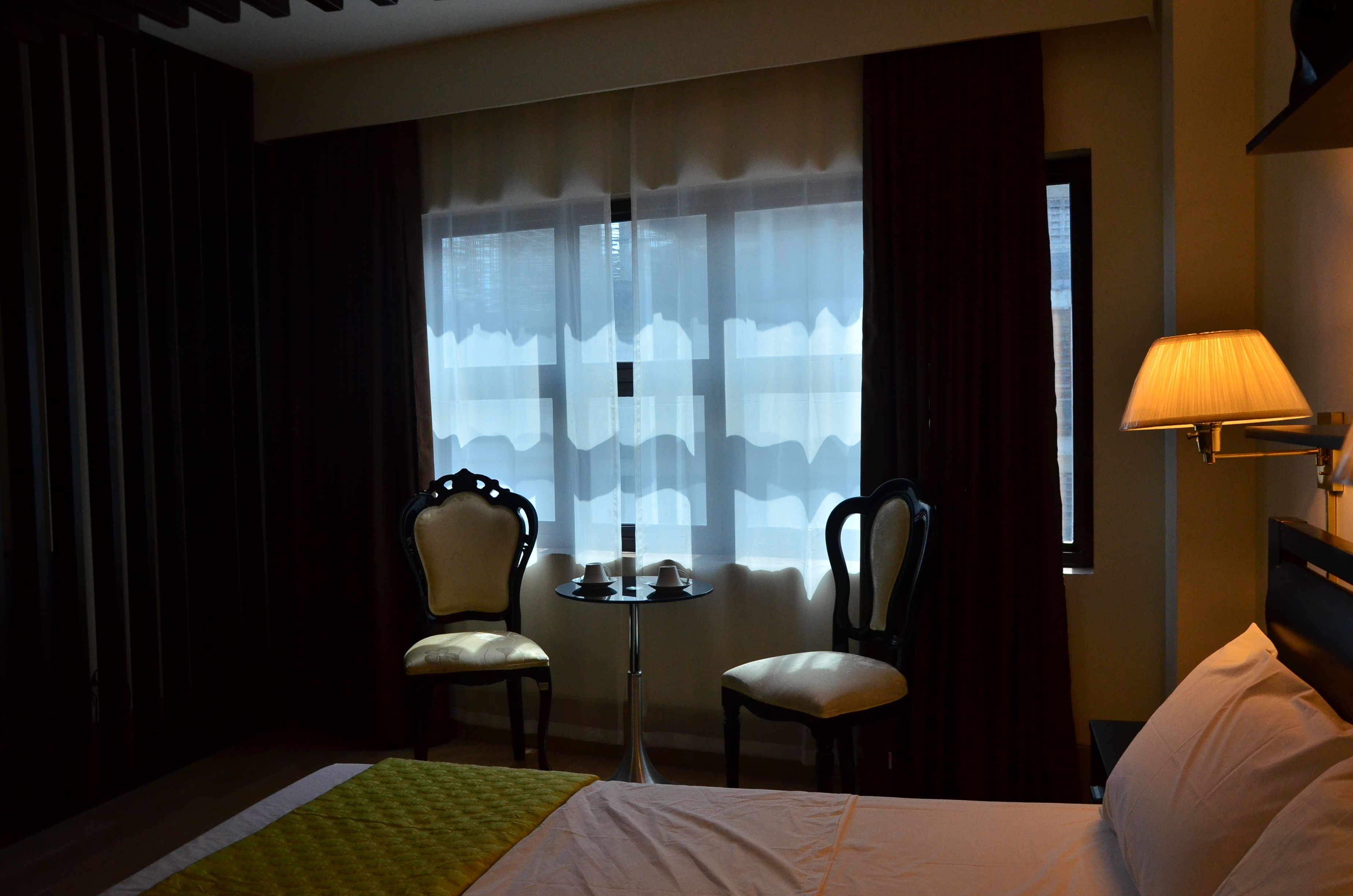 Habitación de hotel con cama, dos sillas, una mesita con tazas, lámpara y ventana con cortinas traslúcidas.