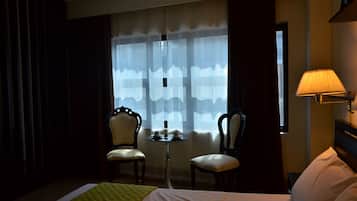 Habitación de hotel con cama, dos sillas, una mesita con tazas, lámpara y ventana con cortinas traslúcidas.