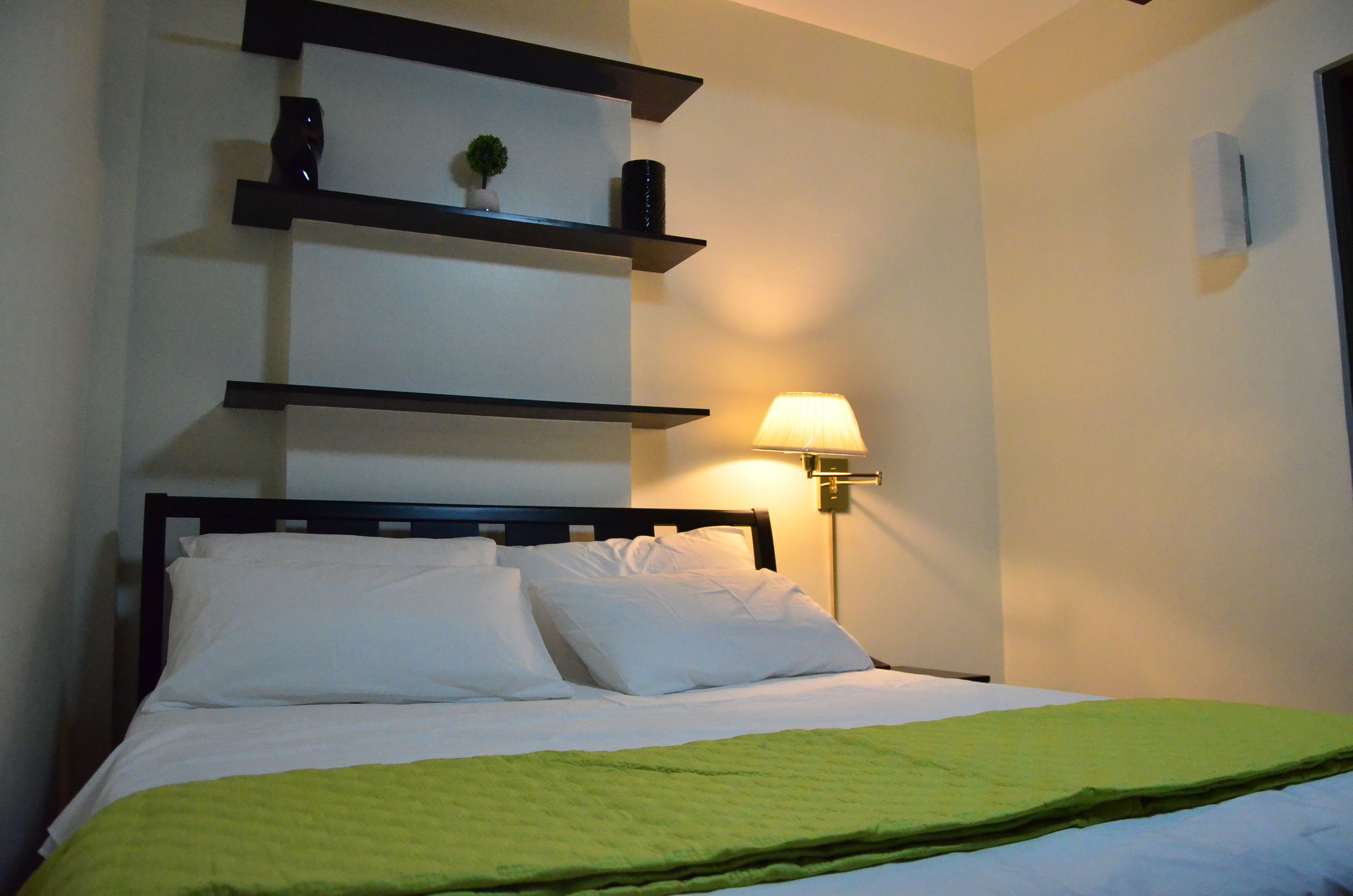 A neatly made bed with white linens and a green blanket, a wall-mounted shelf with decorative items, and a bedside lamp.