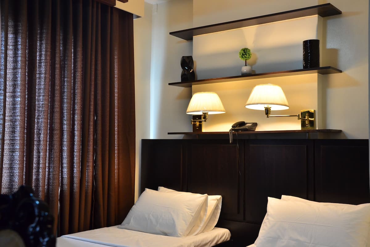 Une chambre d’hôtel avec une tête de lit sombre, deux lampes de chevet, une étagère avec des objets décoratifs et une fenêtre avec des rideaux.