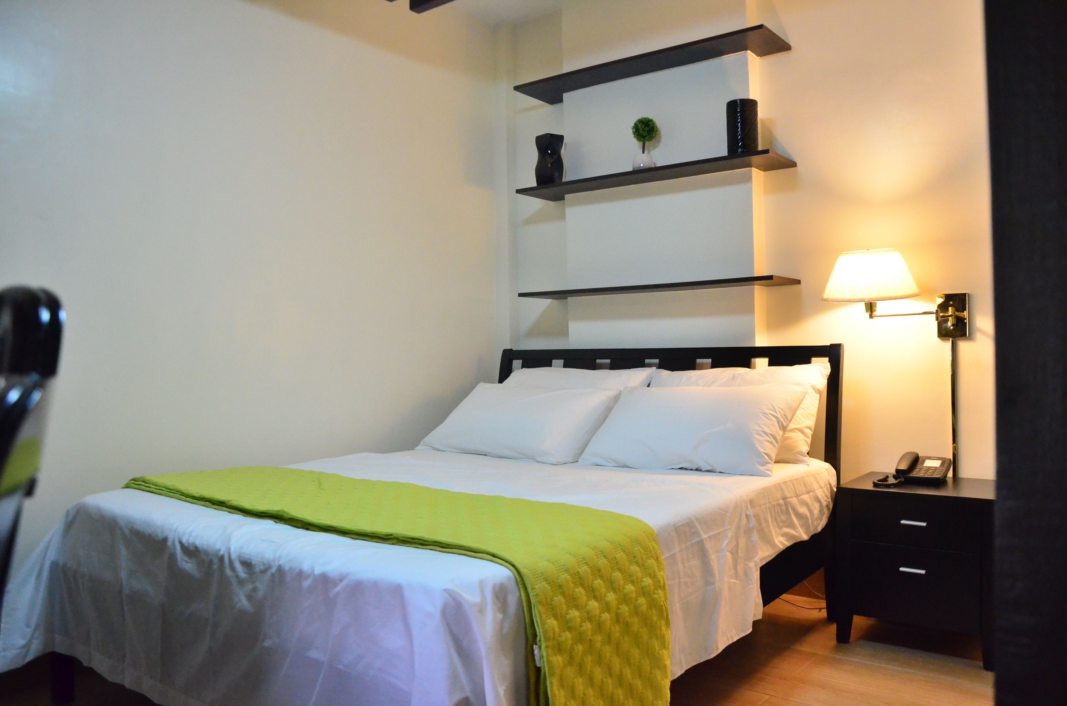Een opgemaakt bed met witte lakens en een groene plaid, een nachtkastje met een lamp en een plank boven het bed.