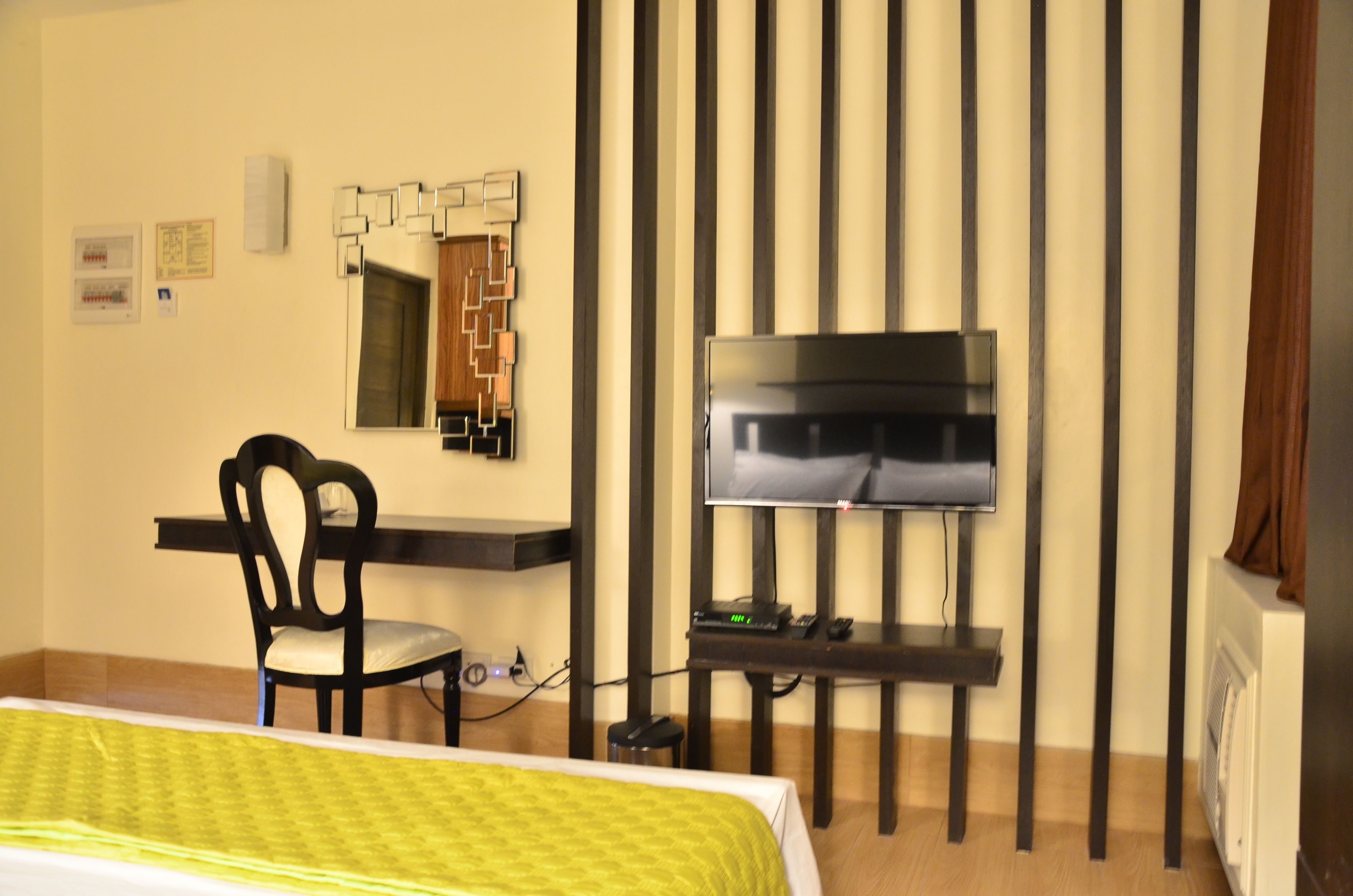 A hotel room with a bed, a desk, a chair, a TV, a mirror, and a wall-mounted heater.