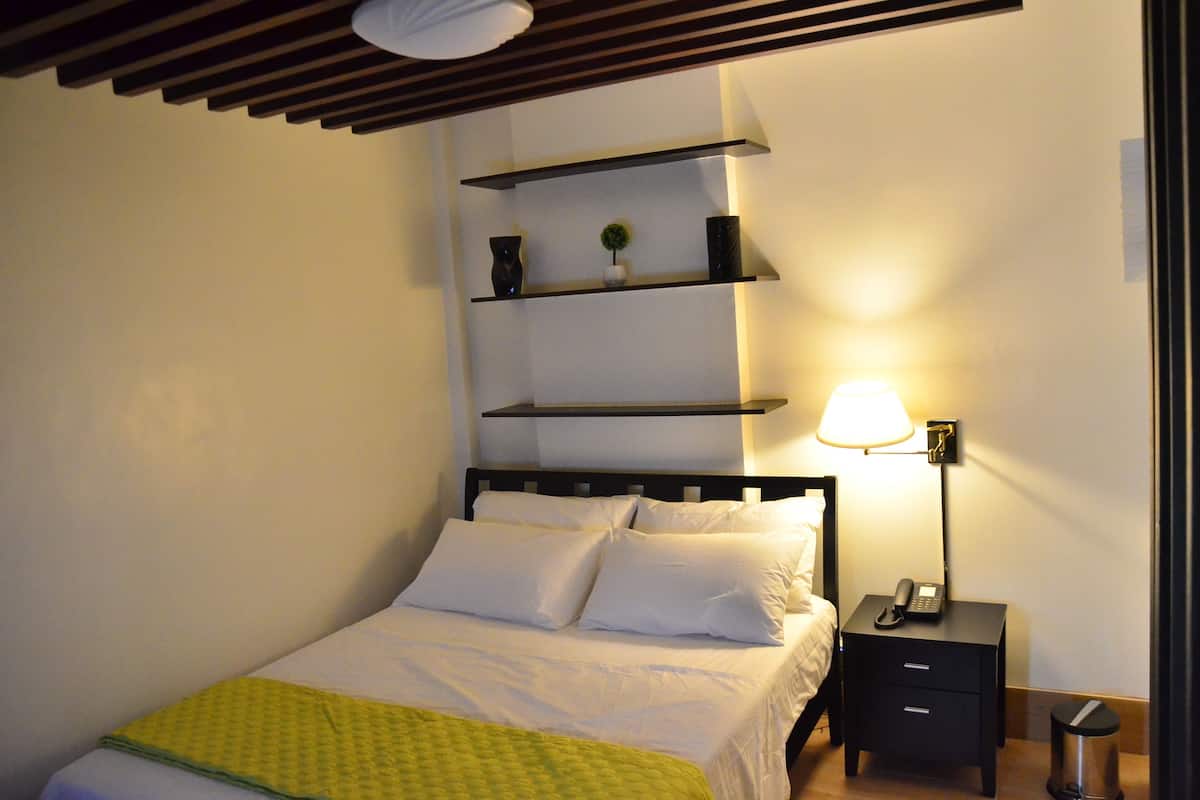 Un lit bien fait, avec du linge de lit blanc et une couverture jaune, une table de chevet avec une lampe et une poubelle.